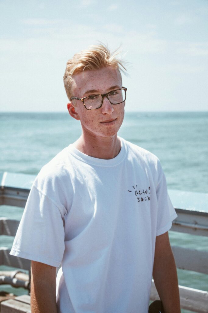 pexels-photo-2589653-2589653 Young man in glasses wearing a white shirt, standing by the sea on a sunny day.